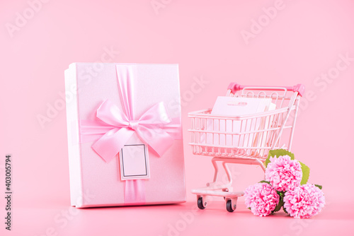 Shopping cart and gift with carnation on pink table background.