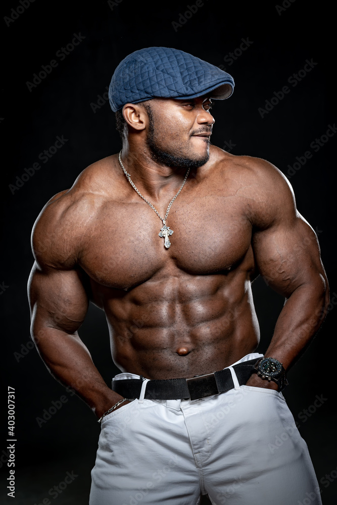 Sexy afro american bodybuilder with no shirt. Athlete poses to the camera in white jeans and blue cap. Strong muscular body.