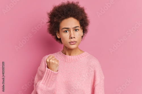 Young Afro American woman shows fist has annoyed face expression going to revenge or threaten someone makes serious look wears casual sweater isolated over pink background. I will show you who is boss