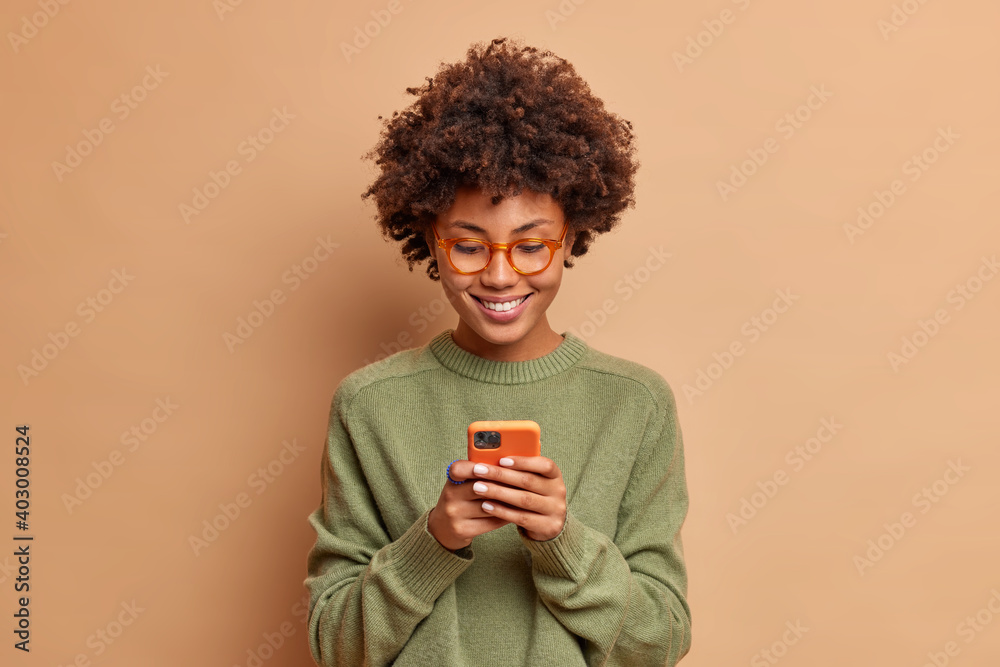 © wayhome.studio - Isolated shot of African American woman uses smartphone application enjoys browsing social media creats news content makes online order wears spectacles and casual jumper poses over beige studio wall © wayhome.studio - Isolated shot of African American woman uses smartphone application enjoys browsing social media creats news content makes online order wears spectacles and casual jumper poses over beige studio wall
