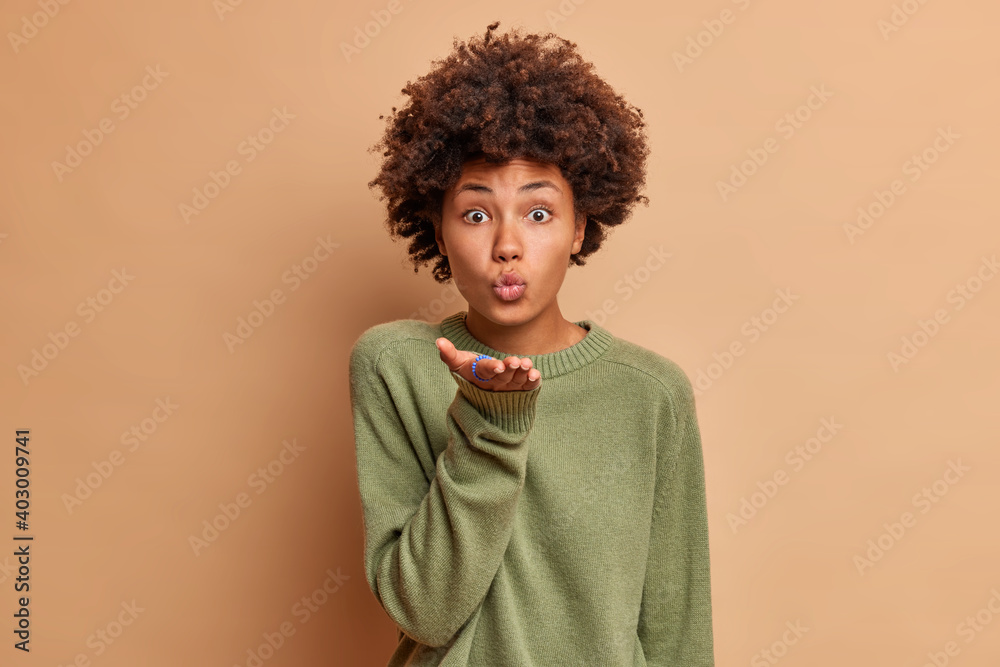 Romantic beautiful African American woman blows air kiss keeps lips folded palm towards mouth sends mwah coquettish wants to cheer up her partner wears casual jumper isolated over beige wall Romantic beautiful African American woman blows air kiss keeps lips folded palm towards mouth sends mwah coquettish wants to cheer up her partner wears casual jumper isolated over beige wall