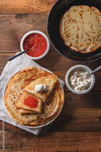 Traditional Russian Crepes Blini stacked in plate and cast-iron frying pan with red caviar, fresh sour cream on dark wooden table. Russian festival meal Maslenitsa or Shrovetide. Top view.