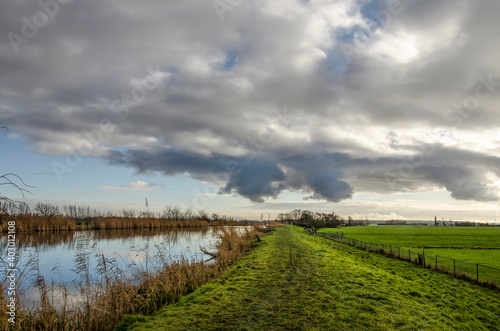 Muddy hiking trail on a grassy dike aling a reeds-lined canal under a dramatic sky on a winter day in vlietlanden nature reserve near Schipluiden, The Netherlands photo