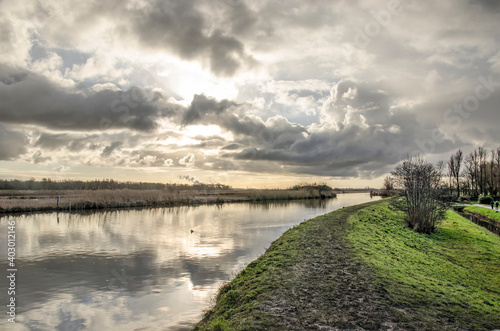 Low sun in a dramatic sky over a muddy hiking trail along a canal on a windless day in winter in Vlietlanden nature reserve near Maasland, The Netherlands
