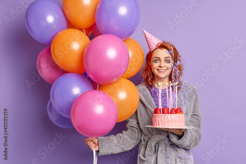 Glad satisfied redhead European woman celebrates special occasion has fun on holiday party poses with strawberry cake wears cone hat inflated colorful balloons isolated over purple background