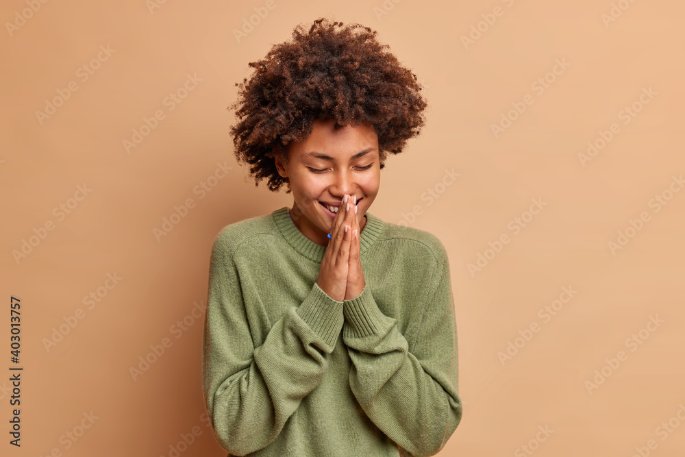 Pleased cheerful dark skinned young woman keeps palms together in pray gesture smiles positively closes eyes has curly hair dressed in casual long sleeved jumper isolated over brown background Pleased cheerful dark skinned young woman keeps palms together in pray gesture smiles positively closes eyes has curly hair dressed in casual long sleeved jumper isolated over brown background