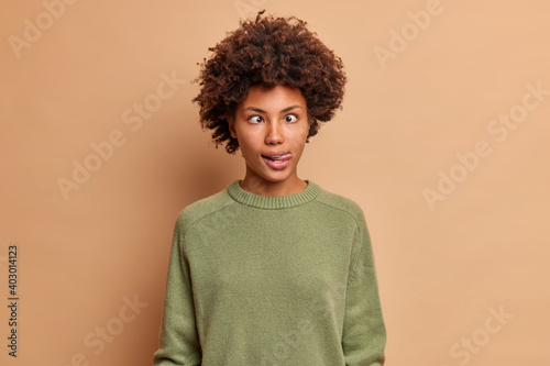 Crazy mad dark skinned woman crosses eyes and sticks out tongue makes grimace foolishes around after all day studying wears casual jumper plays fool isolated over brown background. Funny face