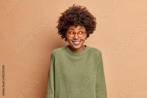 Portrait of pretty positive ethnic woman looks aside with toothy smile sees something nice wears casual long sleeved jumper and spectacles poses in studio against brown background. Good emotions © wayhome.studio 