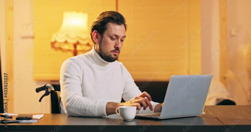 Young handicaped Man Freelancer Using Laptop, Typing, Scrolling, Surfing Web, Looking at the Screen. Professional Creative Millennial Sitting at His Desk in Home Office Studio Working Young handicaped Man Freelancer Using Laptop, Typing, Scrolling, Surfing Web, Looking at the Screen. Professional Creative Millennial Sitting at His Desk in Home Office Studio Working