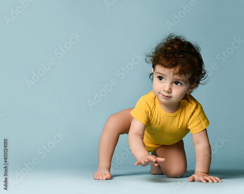 Obraz na płótnie Small cute positive curly baby boy toddler in yellow comfortable jumpsuit crawling on floor and trying to stand up over blue wall background. Happy childhood, stylish look for newborn and children w salonie