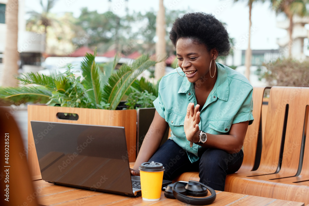 A young African girl sits in a street cafe and communicates with friends and colleagues using a laptop. A beautiful girl is sitting at a table, next to a laptop, coffee, phone and headphones. A young African girl sits in a street cafe and communicates with friends and colleagues using a laptop. A beautiful girl is sitting at a table, next to a laptop, coffee, phone and headphones.