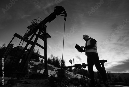 Low angle of petroleum engineer looking at oil pump rocker-machine and writing on clipboard. Silhouette of oil worker checking work of balanced beam petroleum pump jack. Black and white image