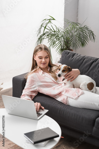 cheerful woman lying on couch with jack russell dog and using laptop in living room © LIGHTFIELD STUDIOS