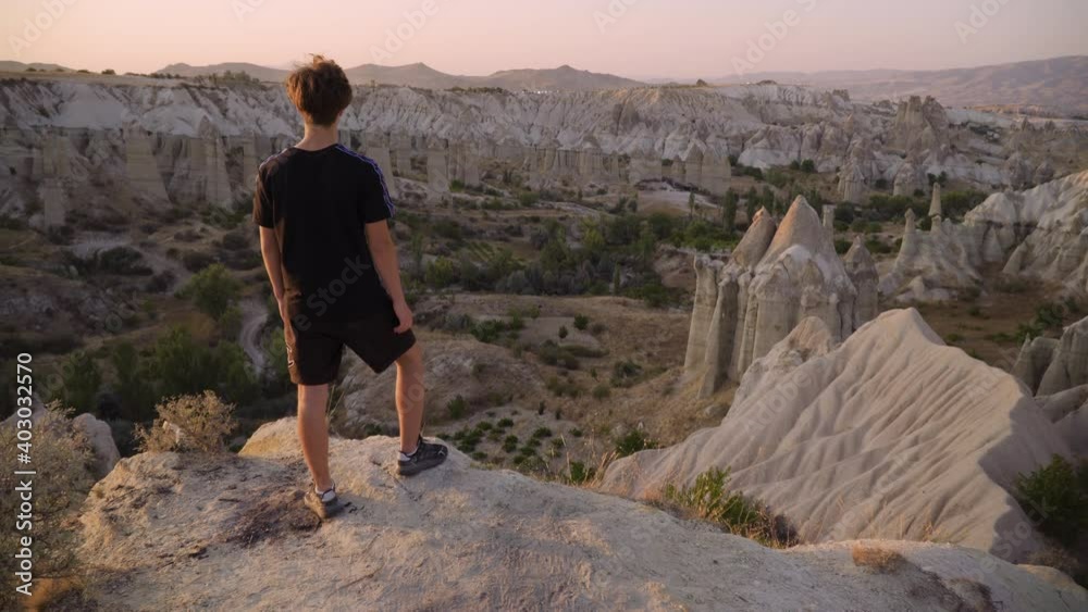 Young man stands on mountain top overlooking view and sunset, Love Valley, Cappadocia, Turkey Young man stands on mountain top overlooking view and sunset, Love Valley, Cappadocia, Turkey