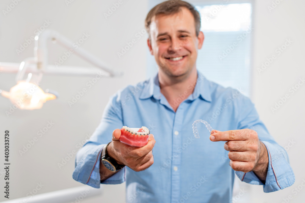Dental care.Smiling orthodontist doctor holding aligners and braces in hand shows the difference between them