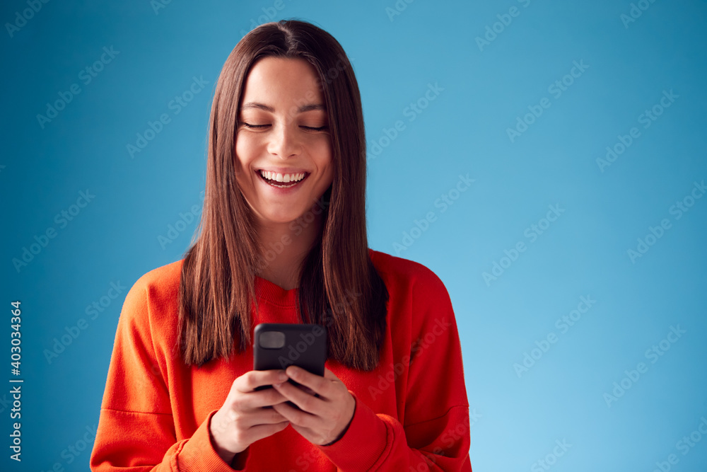 Studio Portrait Of Smiling Young Woman Looking At Mobile Phone Against Blue Background - obrazy, fototapety, plakaty 