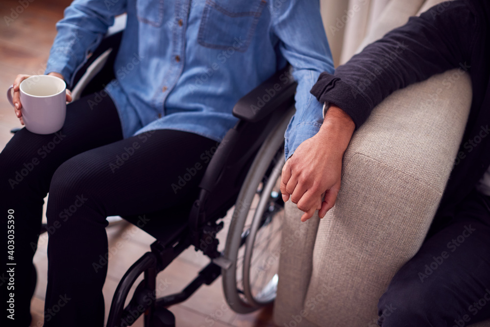 Obraz premium Close Up Of Couple At Home With Disabled Woman In Wheelchair Holding Hands With Man Sitting On Sofa