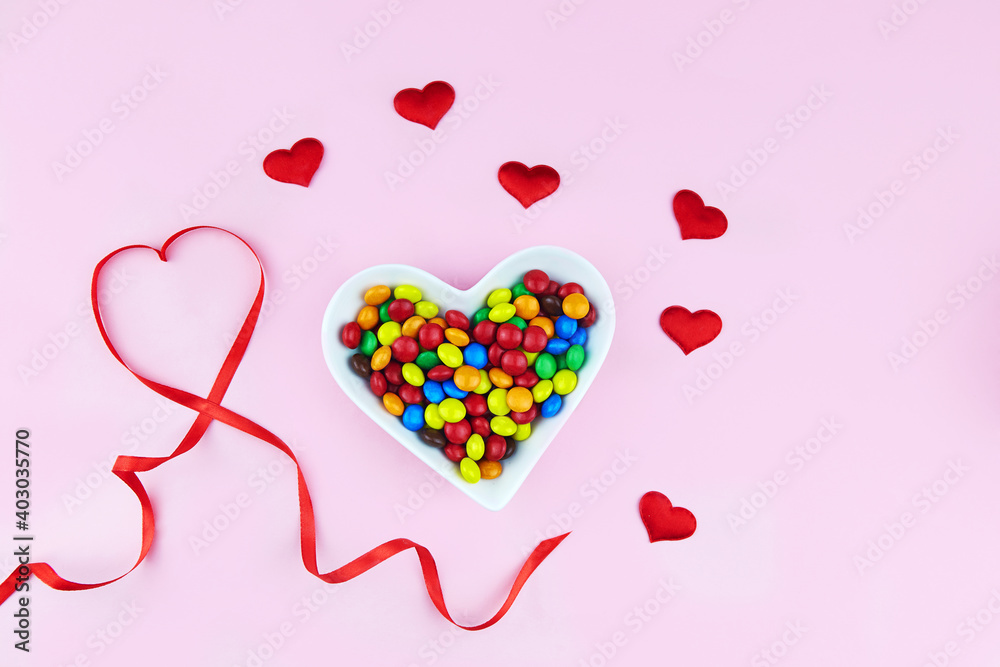 Colorful candies in a heart-shaped plate, red ribbon, hearts and decorations on a pink background. Valentine's Day, Mother's Day, Birthday.