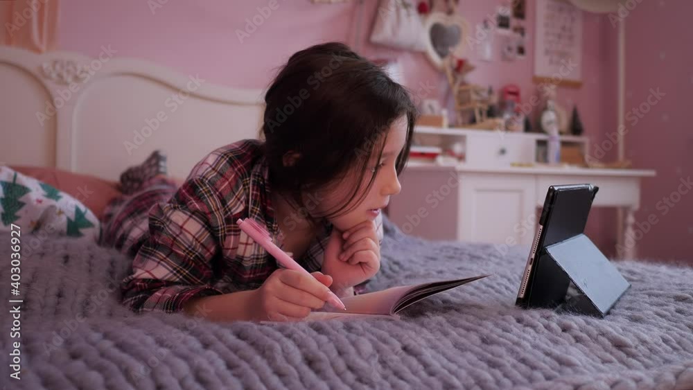 Distance education at home, online e-learning. Cute child is looking in laptop screen lying on bed and writing in notebook. Little school girl pupil doing her homework in school online. Distance education at home, online e-learning. Cute child is looking in laptop screen lying on bed and writing in notebook. Little school girl pupil doing her homework in school online.