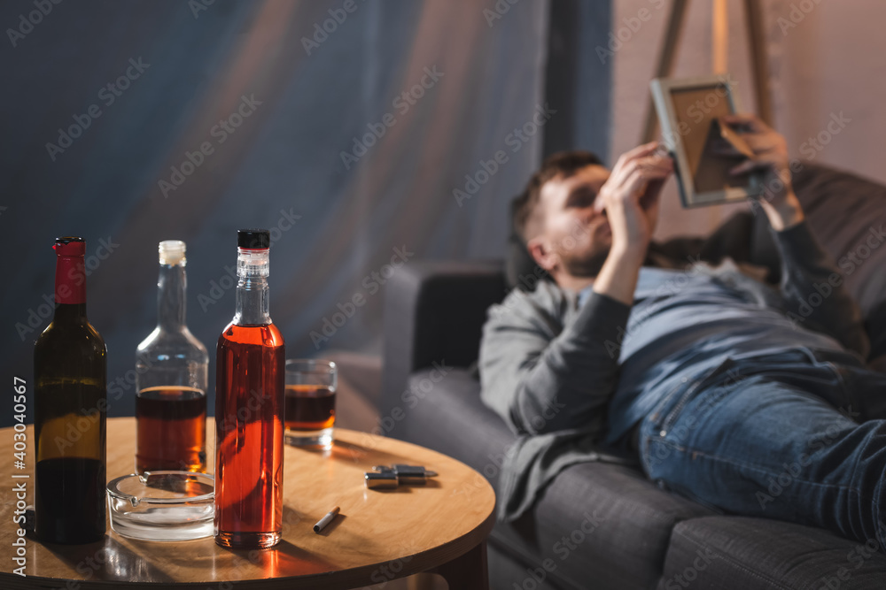 table with bottles of alcohol near depressed man lying on sofa with photo frame on blurred foreground table with bottles of alcohol near depressed man lying on sofa with photo frame on blurred foreground
