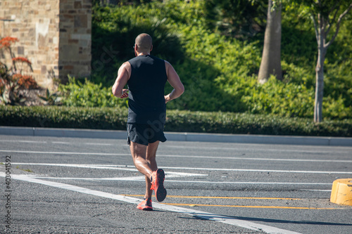 Male runner with shaved head in black tank top jogging in low angle sunlight accross street