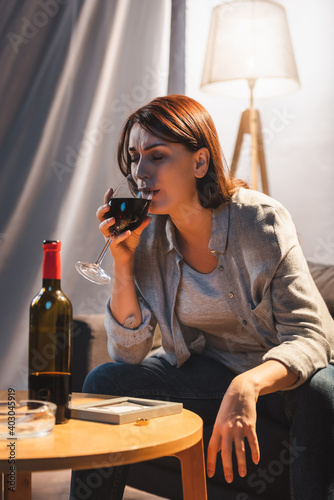 frustrated woman crying and drinking wine while sitting near photo frame and wedding ring on table