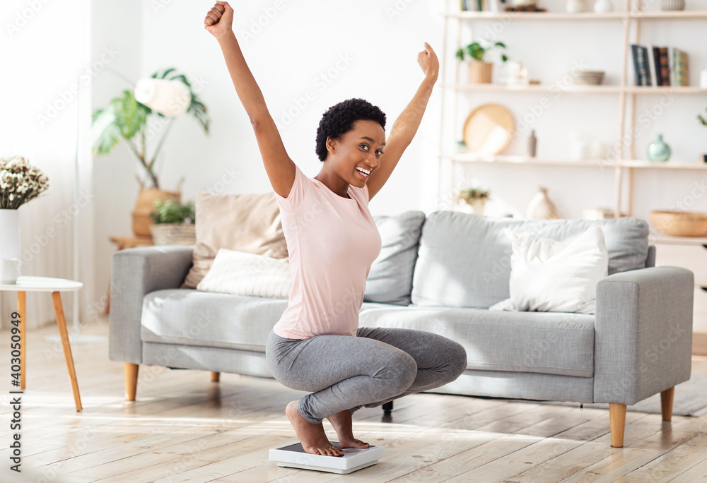 Young black woman sitting on scales, raising hands up in excitement, happy with result of her weight loss diet at home Young black woman sitting on scales, raising hands up in excitement, happy with result of her weight loss diet at home