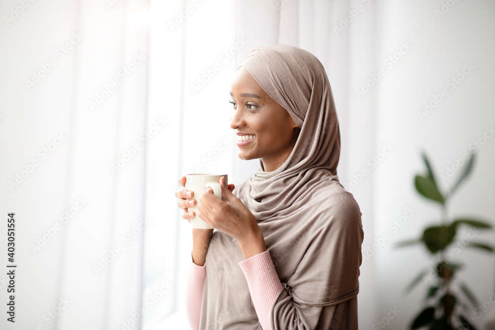 Attractive black woman in hijab with cup of hot coffee looking out window at home, enjoying her hot drink - obrazy, fototapety, plakaty 