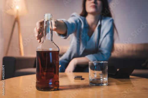 cropped view of alcoholic woman taking bottle of whiskey from table, blurred background photo