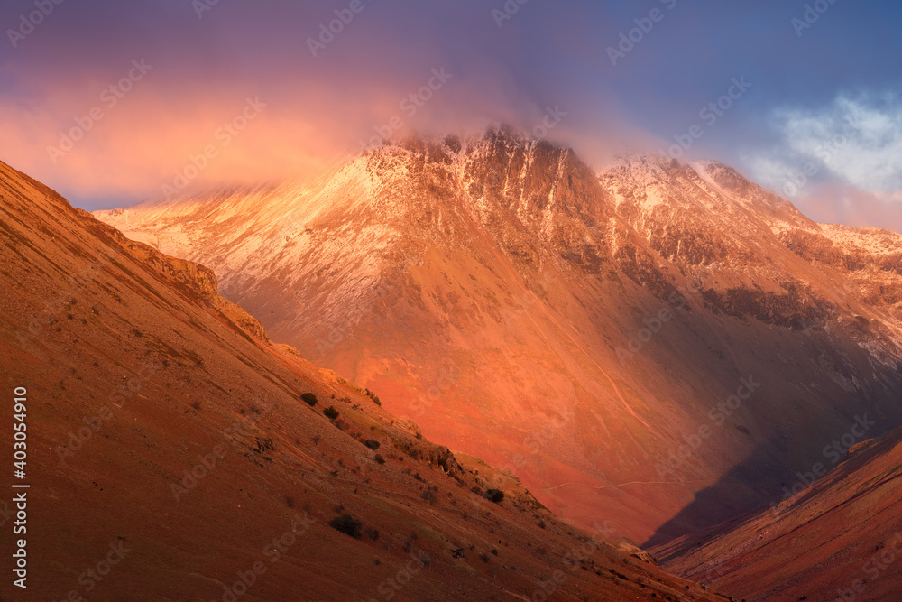 Majestical snowcapped mountains with dark dramatic clouds and red Winter light. Nature background. Lake District National Park, UK.
