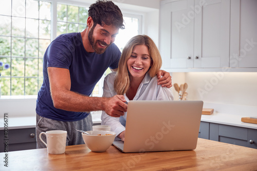Couple Wearing Pyjamas In Kitchen Watching Laptop Whilst Eating Breakfast