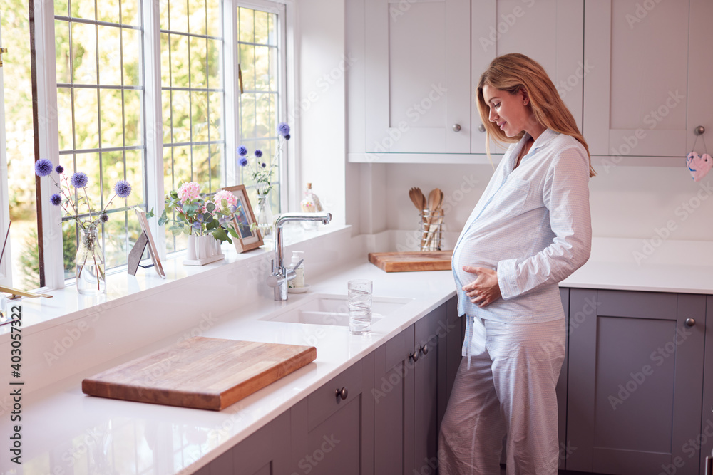 Pregnant Woman Wearing Pyjamas Standing In Kitchen Holding Bump Pregnant Woman Wearing Pyjamas Standing In Kitchen Holding Bump