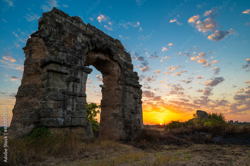Park of the aqueducts ruins of the Claudius aqueduct, Via Appia Antica. Perspective arch during sunset, play of clouds, blue sky, yellow-orange sun reflections on the horizon. Rome, Italy. Appio latin