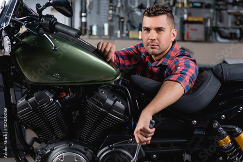 young mechanic looking at camera near motorcycle in workshop