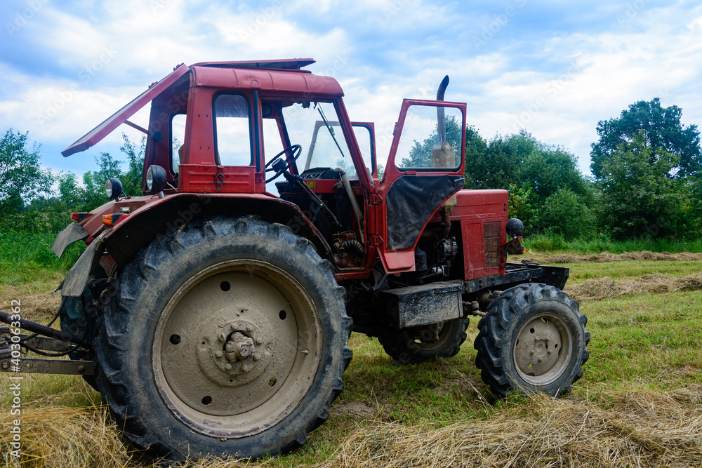 Old red tractor in the field during the haymaking season, pressing hay on bales, forage harvesting. Old red tractor in the field during the haymaking season, pressing hay on bales, forage harvesting.