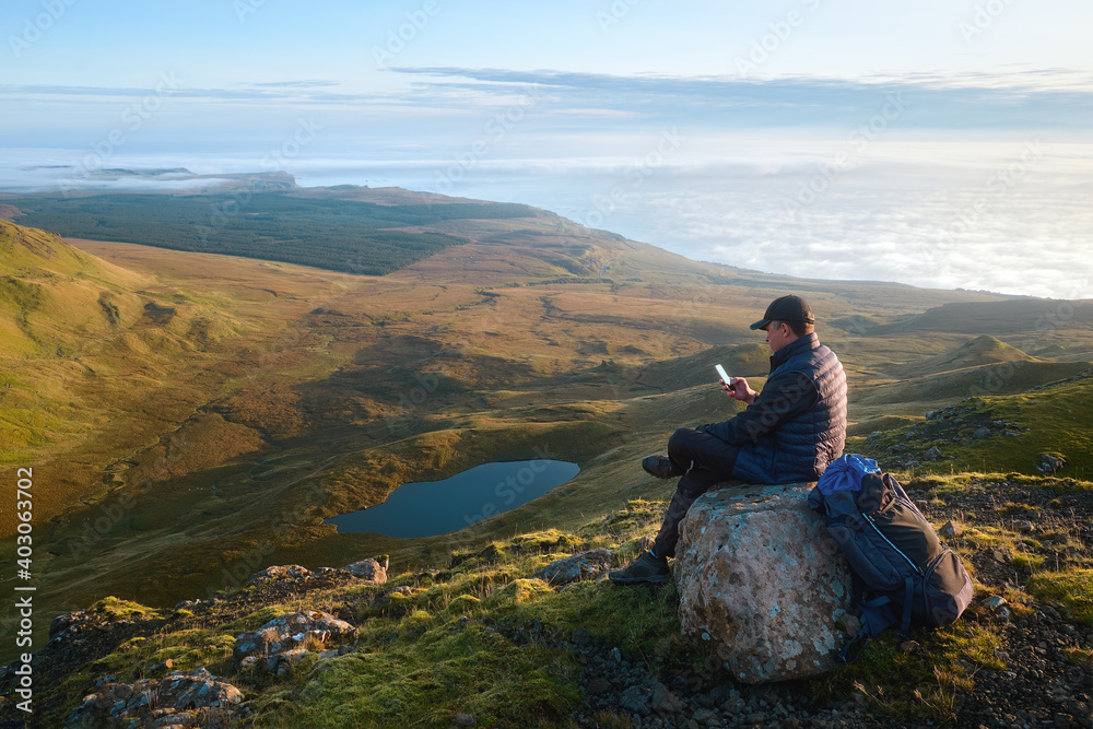 Hiker man sitting on the big stone on the top of the mountain and using his phone at the background of morning landscape with mountains, lake and sea covered clouds. Isle of Skye, Scotland