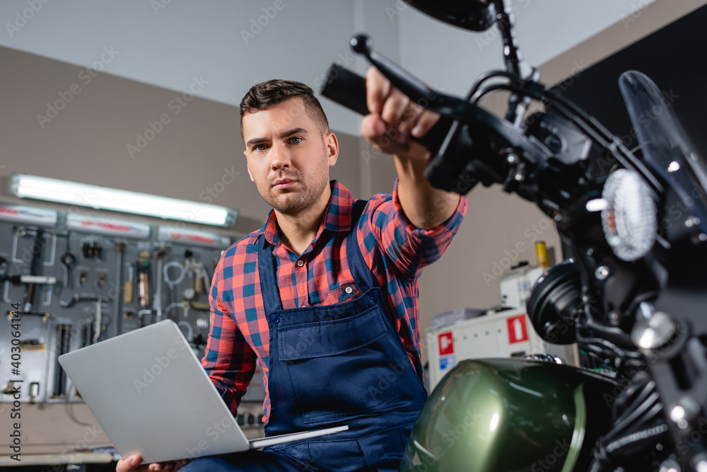 young mechanic with laptop looking at camera while sitting on motorcycle in workshop, blurred foreground