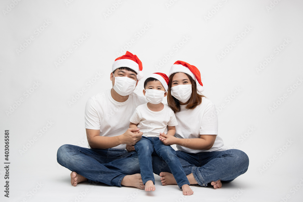 Asian family wearing protective medical mask for prevent virus Covid-19 and wearing christmas hat and sitting together on floor isolated white background Asian family wearing protective medical mask for prevent virus Covid-19 and wearing christmas hat and sitting together on floor isolated white background