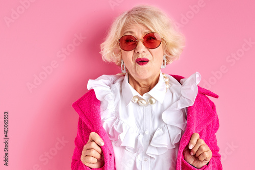 amazed elderly female in stylish clothes posing at camera isolated on pink background, surprised emotional lady in sunglasses