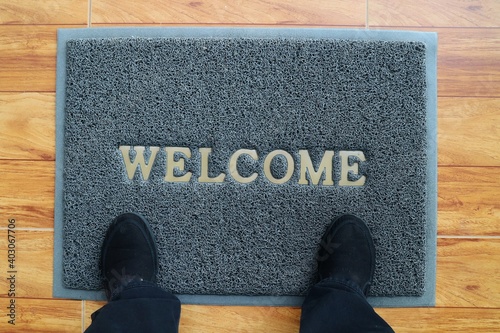 A gray welcome doormat with feet wearing black shoes. Interior and object concept.