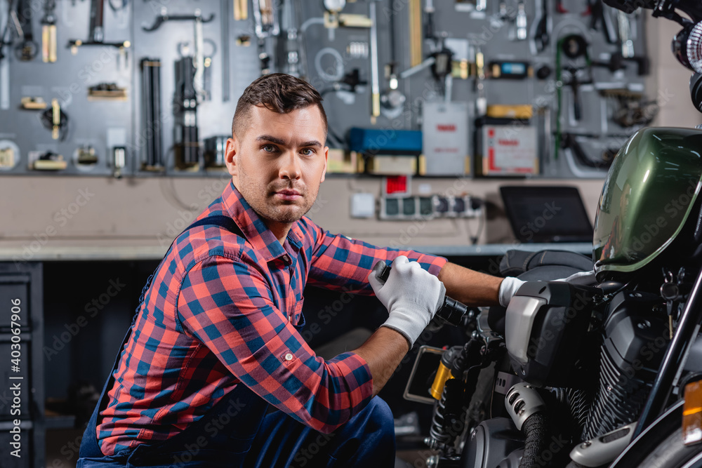 young mechanic looking at camera while examining motorcycle with flashlight young mechanic looking at camera while examining motorcycle with flashlight