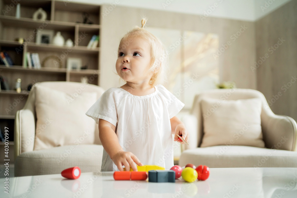 beautiful baby girl playing at home alone with her children's colored wooden toys for development. Blonde of European appearance in a bright apartment on the table in the living room beautiful baby girl playing at home alone with her children's colored wooden toys for development. Blonde of European appearance in a bright apartment on the table in the living room