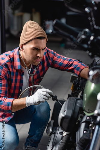 mechanic in plaid shirt and gloves checking engine of motorcycle with stethoscope on blurred foreground