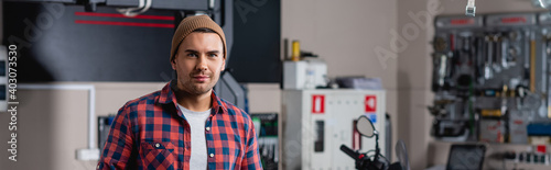 young mechanic in plaid shirt and beanie smiling at camera in workshop, banner