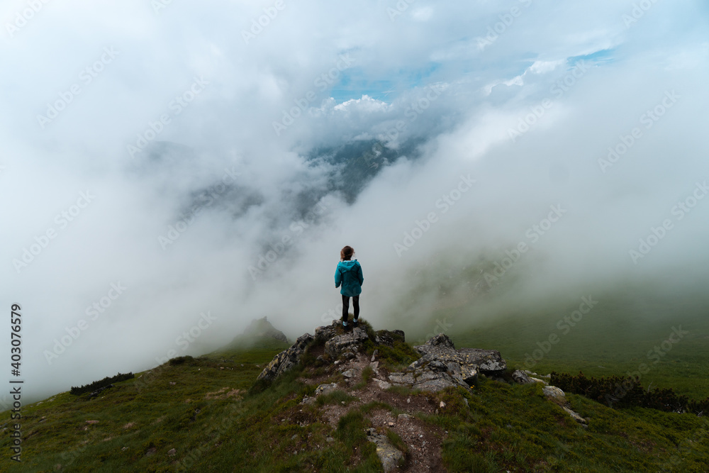 Hiking girl at the edge of a ravine in the Tatra Mountains, on the border between Poland and Slovakia, on a cloudy day with fog and sea of clouds. Blue jacket.