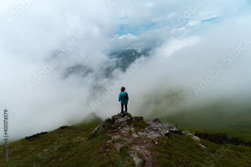 Hiking girl at the edge of a ravine in the Tatra Mountains, on the border between Poland and Slovakia, on a cloudy day with fog and sea of clouds. Blue jacket.