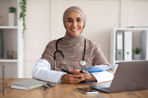 Smiling young woman doctor in hijab taking anamnesis in clinic