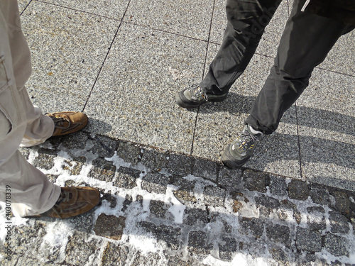 Vier Beine und Stiefel von 2 Geschäftsleuten, die auf grauem Kopfsteinpflaster auf dem Bürgersteig stehen. Die Sonne macht lange Schatten auf dem steinigen Boden. Diese Szene stammt aus Chemnitz photo