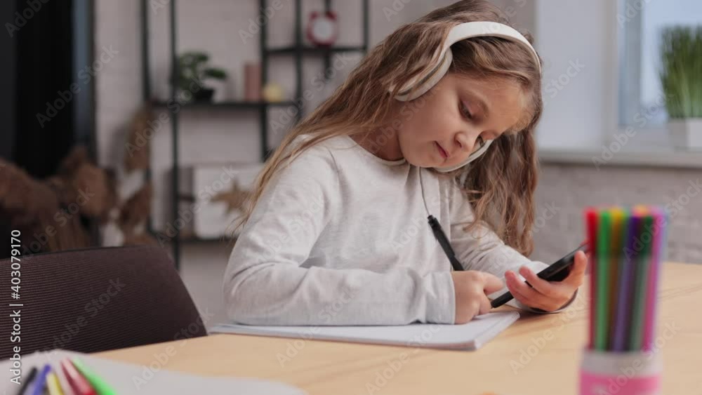 Cute primary school girl in earphones studying at home using smartphone. Schoolgirl has online lesson, writing in exercise book, doing homework. Distance learning online. Children home education Cute primary school girl in earphones studying at home using smartphone. Schoolgirl has online lesson, writing in exercise book, doing homework. Distance learning online. Children home education