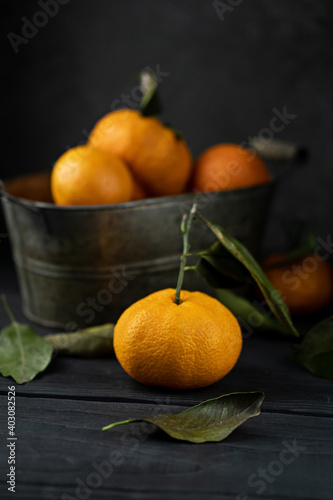 Several ripe tangerines with leaves in a metal container lie on a black table against a dark background. Rustic style with copyspace.